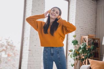 Cheerful young brunette woman in a bright orange sweater enjoying music with headphones indoors in...
