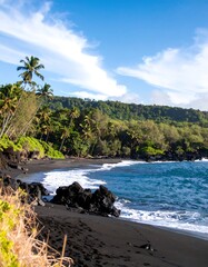 A tranquil, tropical black sand beach stretches along a lush, green hillside under a vibrant blue sky.