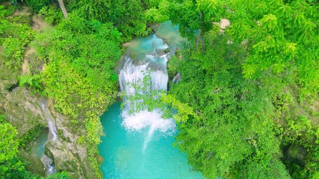 Aerial view of a turquoise waterfall cascading into natural pools, with young men and women swimming, surrounded by green jungle at Kawasan Falls, Cebu, Philippines.
