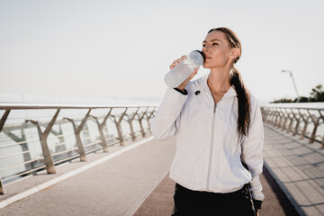 Fit European woman in sportwear relaxing and drinking water on seaside promenade after running and...