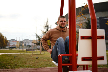 Man doing sit-ups on a red fitness bench outdoors, wearing a hoodie and jeans, showing a strained face while exercising in the park. The concept of a healthy lifestyle