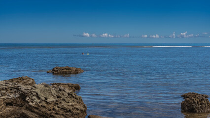 A calm blue ocean. Ripples on the water. Foam waves in the distance. A man in sunglasses is seen floating on his back. Boulders in the foreground. Azure sky, clouds. Madagascar. Nosy Be  