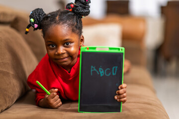 African girl holds a digital drawing tablet with the alphabets ABCD written on the screen, lying on a brown couch at home.