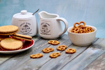 Rustic ceramic milk jug and sugar bowl with cookies, biscuits, and pretzels on wooden table, creating a cozy farmhouse tea time setting with vintage kitchenware and homemade snacks.