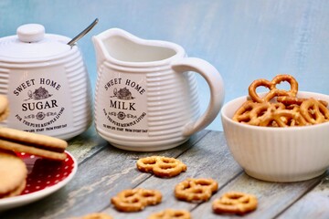 Rustic ceramic milk jug and sugar bowl with cookies, biscuits, and pretzels on wooden table, creating a cozy farmhouse tea time setting with vintage kitchenware and homemade snacks.