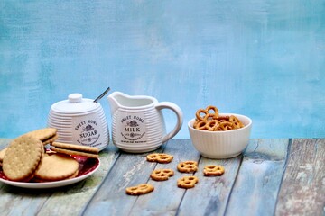 Rustic ceramic milk jug and sugar bowl with cookies, biscuits, and pretzels on wooden table, creating a cozy farmhouse tea time setting with vintage kitchenware and homemade snacks.