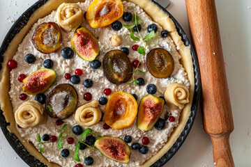 A tart or cottage cheese pie with fruits and berries in a mold is prepared for baking and a rolling pin on a light background or table. View from above. High quality photo