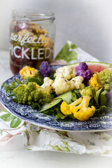 Fresh salad with colorful cauliflower florets, romanesco, cucumber, and greens on a blue plate with a jar of homemade pickled vegetables in the background healthy vegetarian meal with vibrant presence