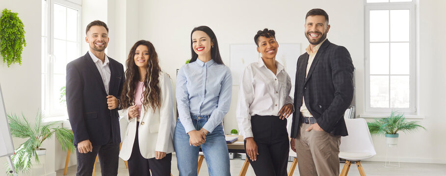 Group of smiling diverse business professionals standing together in modern office space. Successful team of young colleagues in formal and casual wear posing for photo and looking at camera. Banner.