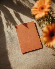Blank orange textured card mockup with blurred gerbera flowers on a sunlit beige textile background. Creative flat lay with deep shadows for branding and creative projects