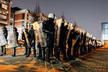 Riot police stand ready in formation during nighttime conflict in an urban area