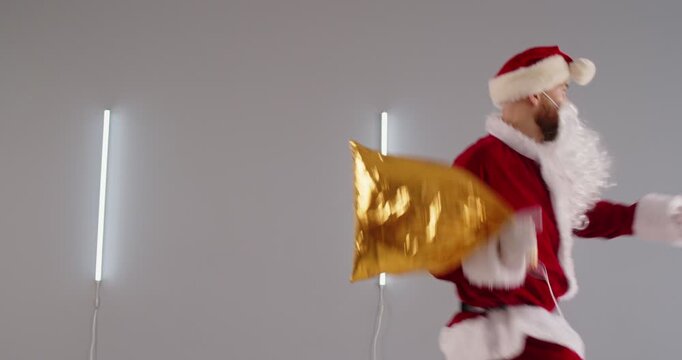 Santa Claus carrying Christmas gold gift bag. Funky confident man with beard, hat and red Xmas costume walking along studio wall with tube LED lights, stumbling on slippery floor and falling backwards