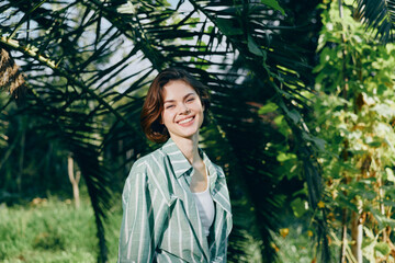 Cheerful woman stands outdoors amid lush greenery, smiling warmly with a relaxed pose, radiating...