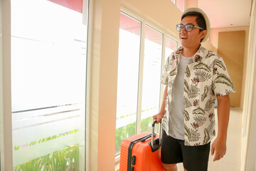 Asian tourist with an orange suitcase, walking through a hotel corridor with large glass windows. Bright atmosphere suitable for concepts of tourism, modern hotel stay, and happy vacation moments.