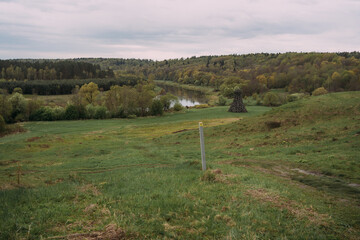 Wide riverside meadow with rolling hills, distant woven twig sculpture and forest under overcast sky