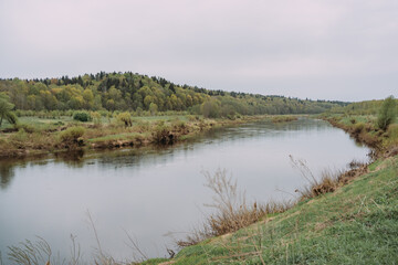 Serene river landscape with calm water reflecting cloudy sky, lush green banks, distant forest and peaceful rural nature in spring, tranquil countryside scene Russia