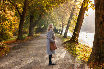 Fototapeta premium Fashion woman in houndstooth coat standing on path in autumn park alley, enjoying sunshine and fall season outdoors