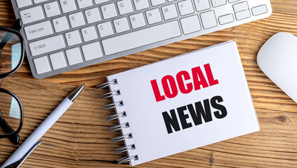 Local news phrase on spiral notebook with keyboard, pen, mouse and glasses on wooden desk