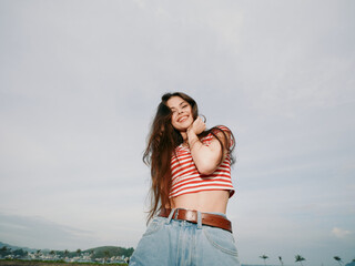 A joyful young woman stands outdoors under a bright sky, wearing a red and white striped crop top...