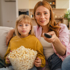 Mother and daughter watching television and eating popcorn on sofa
