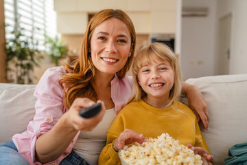 Mother and daughter watching television and eating popcorn on sofa