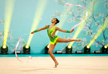 Rhythmic gymnast performing ribbon routine in colorful leotard during artistic gymnastics competition indoors with floral background and stage lighting