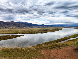 The scene features a vast river landscape, framed by gently rolling hills that stretch into the distance. Above, a dramatic sky filled with vibrant clouds