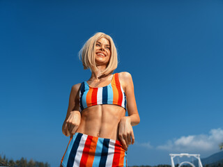 happy young woman wearing colorful striped bikini smiling outdoors, enjoying sunny day with clear blue sky, summer vacation and beach lifestyle concept