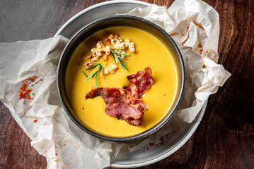 Rustic bowl of creamy yellow soup garnished with crispy bacon, rosemary, and roasted nuts, served on parchment over white plate on dark wooden table. Gourmet autumn comfort food.