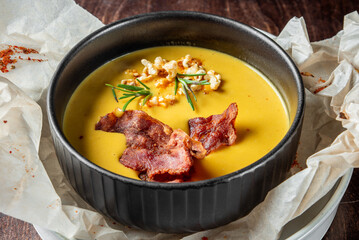 Rustic bowl of creamy yellow soup garnished with crispy bacon, rosemary, and roasted nuts, served on parchment over white plate on dark wooden table. Gourmet autumn comfort food.