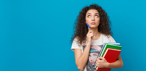 Thoughtful smart female student holding pen and having an idea, looking up pensive, carry notebooks, standing against blue background