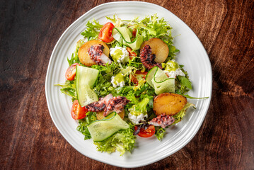 Gourmet salad with grilled octopus, roasted potatoes, cherry tomatoes, cucumber ribbons, leafy greens, and creamy cheese, served on a white plate over dark wooden background.