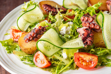 Gourmet salad with grilled octopus, roasted potatoes, cherry tomatoes, cucumber ribbons, leafy greens, and creamy cheese, served on a white plate over dark wooden background.