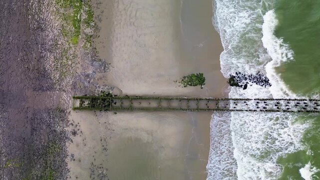 Aerial view of ocean waves crashing against rows of stone breakwaters. The geometric pattern of the structures contrasts with the dynamic movement of the sea, creating a striking coastal scene.