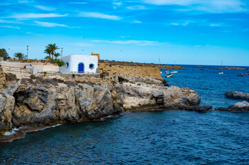 A rocky shoreline with a small white building and a blue roof. The water is calm and the sky is...