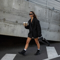 Fashionable young woman in oversized black blazer and sunglasses holding takeaway coffee cups,...