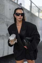 Stylish young woman in oversized black blazer and sunglasses walking outdoors with takeaway coffee...