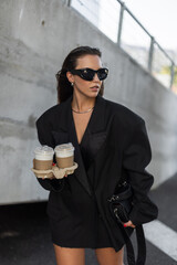 Trendy female in street style outfit, oversized blazer and accessories, holding coffee cups, urban...