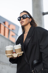 Stylish young female in trendy oversized blazer holding coffee to go, wearing black accessories and...
