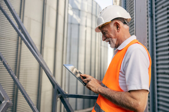 Side view of a senior heavy industry worker in protective wear standing against the silo and scrolling on tablet.
