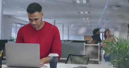 Sitting man in red shirt typing at laptop in open-plan office with coffee-cup black folder plant