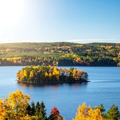 Obraz premium A serene autumnal vista showcases a tranquil lake, dotted with an island ablaze in vibrant fall colors, amidst a landscape of colorful trees and hills.
