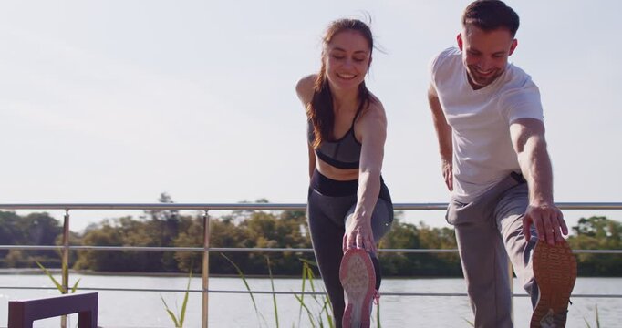 Sports exercises and training of fit couple of athletes before healthy jogging in summer park. Sporty happy woman and man runners in sportswear bend over together to stretch muscles and warm up
