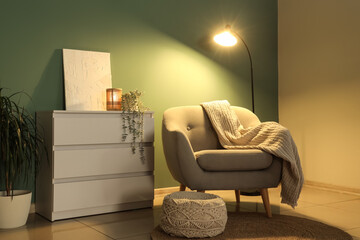 Comfortable armchair, pouf, chest of drawers and glowing floor lamp at night in interior of living...