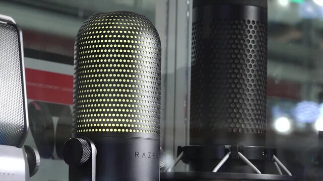 Close-up of high-end gaming and studio microphones on display behind glass, including a Razer mic with illuminated yellow LED pattern and metal body.
