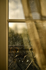 Sunlit raindrops on a windowpane with a soft glimpse of the window frame and sheer curtain, gently blurred. A warm, peaceful scene evoking quiet moments of calm after spring rain. Vertical photo.