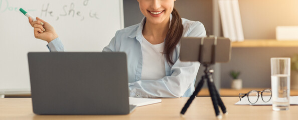 Modern online lessons, tutoring, new normal during covid lockdown. Smiling young arabian lady teacher shows to blackboard and looks at webcam on smartphone on table with laptop in home interior