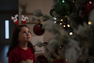 Little girl wearing reindeer antlers looking up at christmas tree