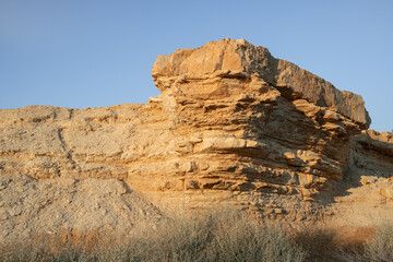 layered rock formation of ochre-colored stone rises from the Negev desert landscape. The eroded geological structure is set against a clear sky with sparse vegetation. Israel south