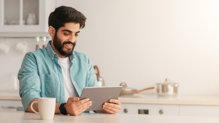 Cheerful arab man using digital tablet while drinking coffee in kitchen at home, sitting at table, enjoying morning beverage and checking social networks on tablet computer, copy space
